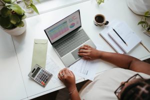 a woman sitting at a desk with a laptop, calculator, papers, and a notebook